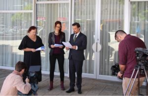 A photograph of 3 people dressed in business attire ,holding a document. They are stand in front of the ACT Legislative Assembly Building, and are being filmed by two news service camera operators. 