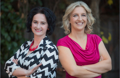 An image of two women standing back to back and smiling at the viewer. One is wearing a bright pink top and the other a zig-zag stripe jacket.
